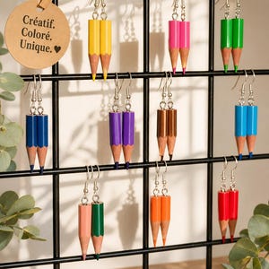 May include: A selection of pencil-shaped earrings in a range of colours, such as yellow, pink, green, blue, purple, brown, orange, and red. The earrings are suspended on a black grid, with a tag that reads "Créatif. Coloré. Unique."