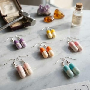 Peut inclure: Boucles d'oreilles artisanales en forme de pilules de différentes couleurs : violet, jaune, orange, rose et bleu, avec des crochets en argent. Les boucles d'oreilles ont de petits visages souriants. L'image comprend également un petit marteau, des cristaux et une bouteille.