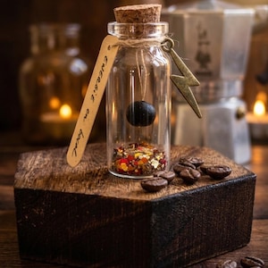 May include: A small glass bottle with a cork stopper, containing a black sphere and colorful glitter. A tag with the text "Energie en boca" hangs from the bottle. A gold lightning bolt charm is attached. The bottle sits on a wooden block with coffee beans.