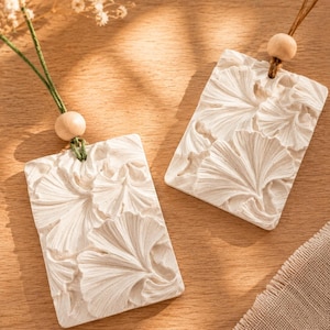 Può includere: Due ornamenti rettangolari in ceramica bianca con un motivo a foglie di ginkgo in rilievo. Ogni pezzo è sospeso da un cordino sottile infilato in un foro superiore, impreziosito da una perlina in legno naturale sopra la superficie ceramica.