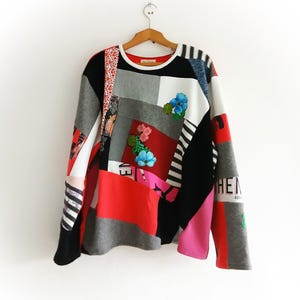 Puede incluir: Una sudadera de manga larga con patchwork y una variedad de colores y patrones. El diseño incluye bloques rojos, grises, negros y blancos, con adornos florales. La manga presenta el texto "HEN".