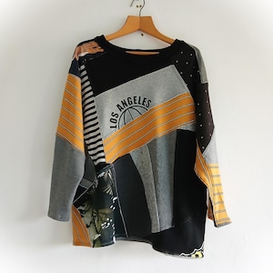 Pode incluir: Uma camisola de manga comprida feita a partir de um patchwork de vários tecidos em preto, cinzento e amarelo mostarda. A peça apresenta uma impressão gráfica de um tigre, padrões às riscas e o texto LOS ANGELES com um ícone de globo.