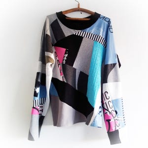 Op de afbeelding: Een patchwork trui met lange mouwen in verschillende kleuren, waaronder grijs, zwart, blauw en roze. De trui heeft een ronde hals en hangt aan een houten kleerhanger. De mouwen hebben tekst- en grafische details.