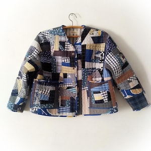 May include: A patchwork jacket with a variety of blue, beige, and white fabric squares. The jacket has two front pockets, a V-neck, and a button closure. It is hanging on a wooden hanger.