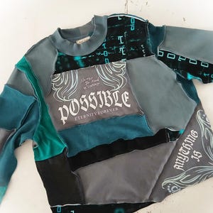 May include: A patchwork sweatshirt in shades of teal, gray, and black. The design incorporates various fabric pieces, with the word "POSSIBLE" and the phrase "Anything is" printed on the front. The sleeves and neckline are a solid teal color.
