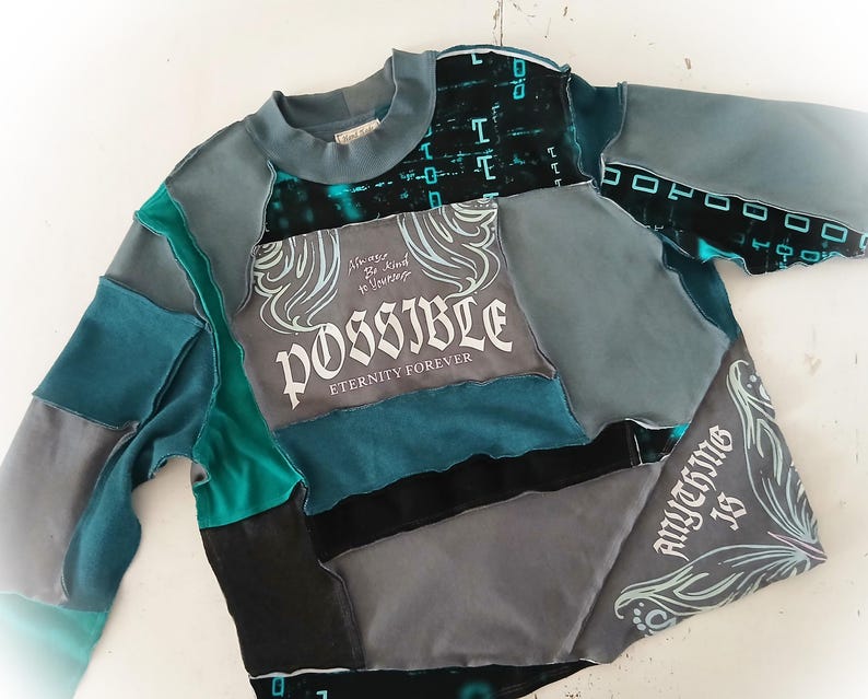 May include: A patchwork sweatshirt in shades of teal, gray, and black. The design incorporates various fabric pieces, with the word "POSSIBLE" and the phrase "Anything is" printed on the front. The sleeves and neckline are a solid teal color.