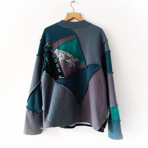 May include: A patchwork sweatshirt in shades of teal, gray, and purple hangs from a wooden hanger. The design features geometric shapes and a high neckline. The sleeves and body are pieced together with visible stitching.