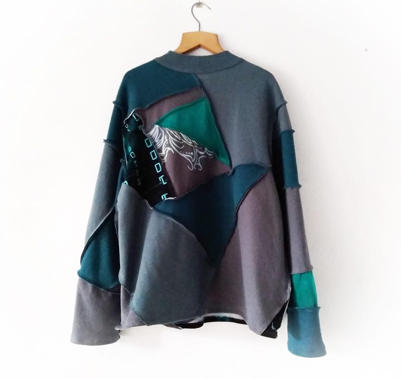 May include: A patchwork sweatshirt in shades of teal, gray, and purple hangs from a wooden hanger. The design features geometric shapes and a high neckline. The sleeves and body are pieced together with visible stitching.