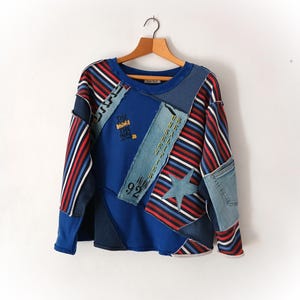 Peut inclure: Un sweat-shirt bleu patchwork avec des détails en denim et en tissu rayé. Le design comprend une étoile, le numéro 92 et le texte "THE MORE THE MOVE". Le haut à manches longues est suspendu à un cintre en bois.