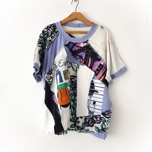May include: A short-sleeved shirt constructed from various fabric panels in light purple, white, and black. The garment features a mix of floral patterns, a graphic of an orange backpack with the text Go Explore, and a surf-themed print with Australia.