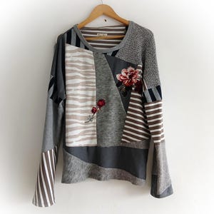 Könnte beinhalten: Ein langärmliges Patchwork-Oberteil mit rundem Ausschnitt. Das Oberteil weist verschiedene Stoffmuster auf, darunter Streifen, Zebramuster und Blumenstickereien. Die Farbpalette umfasst Grau, Braun und Weiß. Das Oberteil hängt an einem Holzbügel.