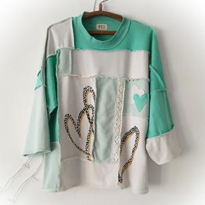 Könnte beinhalten: Ein Patchwork-Langarmshirt in Weiß, Mintgrün und Creme. Das Shirt hat einen Rundhalsausschnitt und ist mit Leopardenherzen, Spitze und einem grünen Herz-Applikation verziert. Die Ärmel sind dreiviertellang.