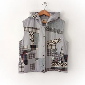 Puede incluir: Un chaleco sin mangas con capucha en gris claro que presenta un diseño de patchwork con varios patrones de tela, incluyendo cuadros, flores y estampados geométricos. El cierre frontal consta de cinco broches de metal plateado sobre una percha de madera.