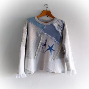 May include: A patchwork sweatshirt in grey, white, and blue, featuring a blue star and lace embellishments. The long-sleeved top hangs on a wooden hanger. The design incorporates various fabric textures and patterns, against a white wall.