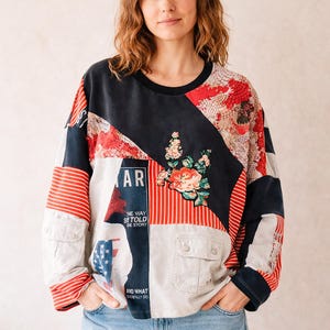 以下が含まれることがあります： 黒、白、赤のパネルを組み合わせた長袖のパッチワークスウェットシャツ。花柄のグラフィック、赤と白のストライプ、アメリカ国旗の一部、そして「AR」、「THE WAY」、「STOLD」、「THE STORY」、「AND WHAT SHE REALLY DID」というテキストがデザインされています。