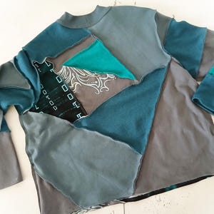 May include: A patchwork long-sleeve shirt in shades of teal, gray, and black. The shirt features a mock neck and is constructed from various fabric pieces sewn together. Some pieces have decorative patterns.