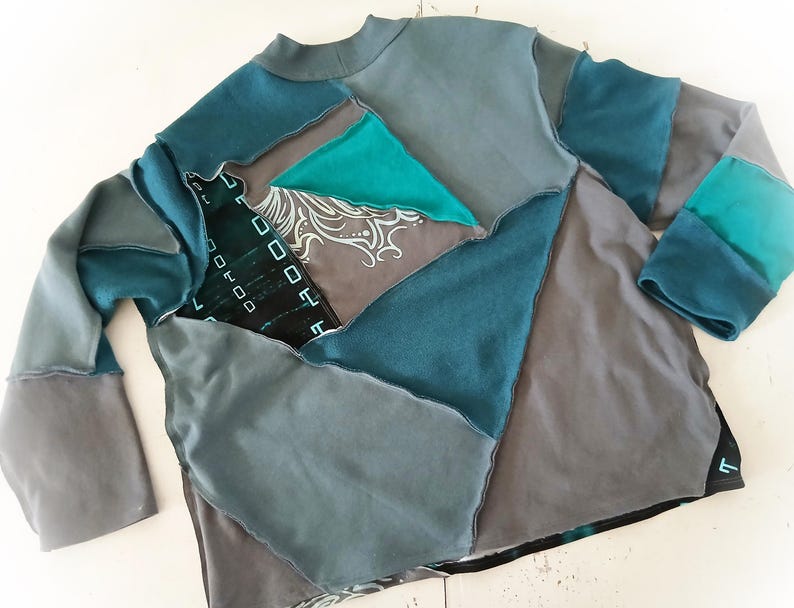May include: A patchwork long-sleeve shirt in shades of teal, gray, and black. The shirt features a mock neck and is constructed from various fabric pieces sewn together. Some pieces have decorative patterns.