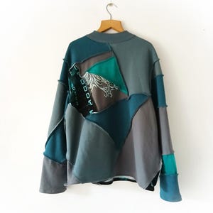 May include: A patchwork sweatshirt in shades of teal, gray, and charcoal hangs on a wooden hanger. The design features geometric shapes and a high neckline. The sleeves are long, and the overall style is casual and unique.