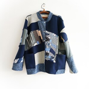 Puede incluir: Una chaqueta patchwork en tonos azules, grises y beige cuelga de una percha de madera. La chaqueta presenta una variedad de patrones y texturas de tela, incluyendo parches con las letras "NW". Tiene una botonadura y dos bolsillos delanteros.