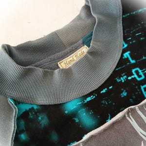 May include: Close-up of a handmade sweatshirt with a gray ribbed collar and a label that reads "Hand Made." The shirt features a teal and black design with a digital pattern and a gray panel with a zebra-like pattern.