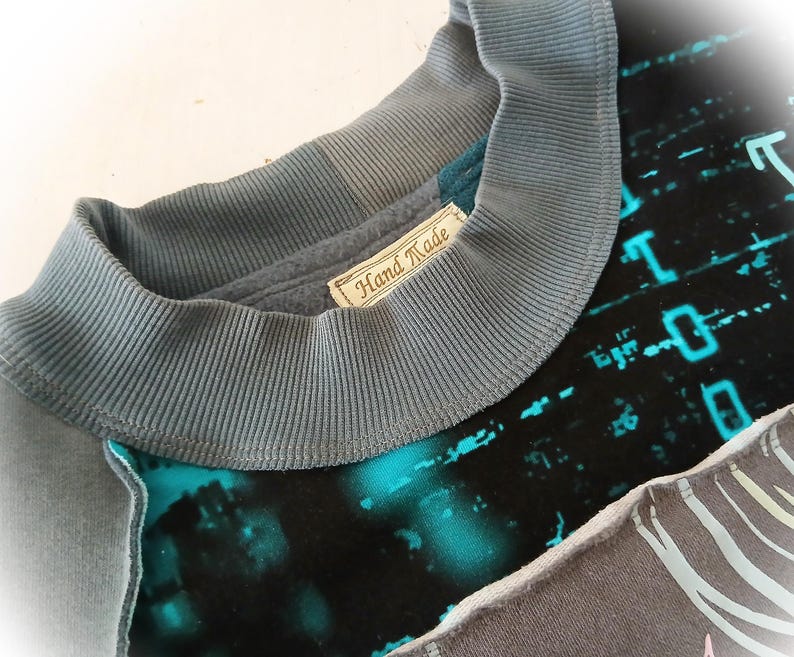 May include: Close-up of a handmade sweatshirt with a gray ribbed collar and a label that reads "Hand Made." The shirt features a teal and black design with a digital pattern and a gray panel with a zebra-like pattern.