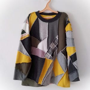 Könnte beinhalten: Ein Patchwork-Langarmshirt mit Rundhalsausschnitt, das an einem pfirsichfarbenen Kleiderbügel hängt. Das Shirt weist eine Vielzahl von Stoffflicken in Grau-, Gelb-, Olivgrün- und Weißtönen auf. Das Design ist eine Mischung aus geometrischen Formen und Texturen.