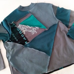 May include: A patchwork long-sleeve shirt in shades of teal, gray, and charcoal. The shirt features a crew neck and is constructed from various fabric pieces, creating a geometric design. Some sections have patterned details.