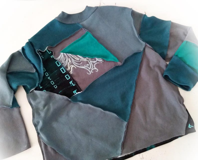 May include: A patchwork long-sleeve shirt in shades of teal, gray, and charcoal. The shirt features a crew neck and is constructed from various fabric pieces, creating a geometric design. Some sections have patterned details.