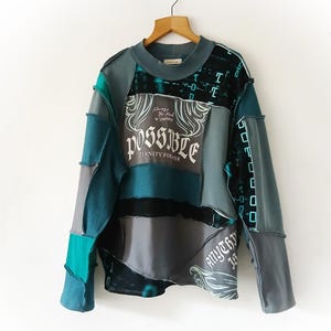 May include: A patchwork long-sleeve shirt in shades of teal, gray, and black. The shirt features the word "POSSIBLE" in a decorative font, along with the phrases "Eternity Forever" and "Anything is". The garment is hanging on a wooden hanger.