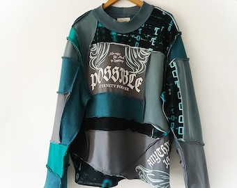 Könnte beinhalten: Ein Patchwork-Langarmshirt in den Farbtönen Teal, Grau und Schwarz. Das Shirt zeigt das Wort "POSSIBLE" in einer dekorativen Schriftart sowie die Sätze "Eternity Forever" und "Anything is". Das Kleidungsstück hängt an einem Holzkleiderbügel.
