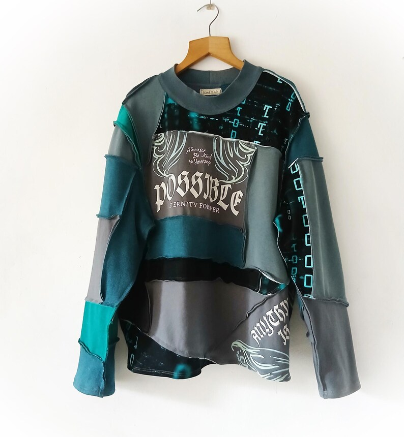May include: A patchwork long-sleeve shirt in shades of teal, gray, and black. The shirt features the word "POSSIBLE" in a decorative font, along with the phrases "Eternity Forever" and "Anything is". The garment is hanging on a wooden hanger.