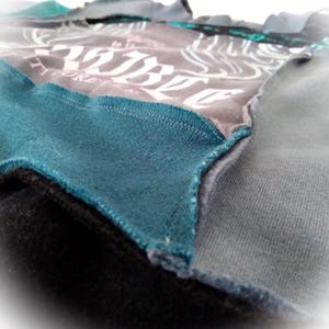 May include: Close-up of a patchwork garment featuring various fabric textures and colors. The design incorporates shades of gray, teal, and black. The gray fabric has a white graphic print. The edges are stitched together, showcasing the construction.
