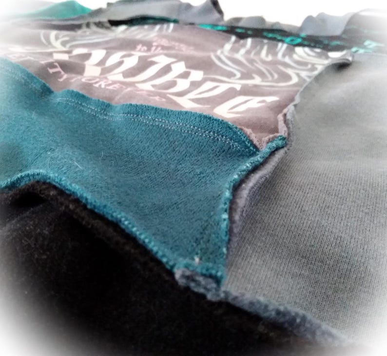 May include: Close-up of a patchwork garment featuring various fabric textures and colors. The design incorporates shades of gray, teal, and black. The gray fabric has a white graphic print. The edges are stitched together, showcasing the construction.