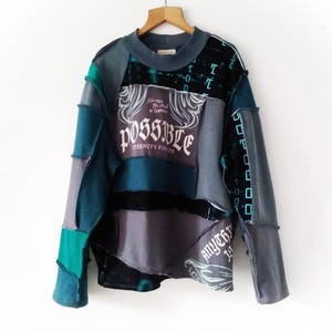 May include: A patchwork sweatshirt in shades of teal, gray, and black, hanging on a wooden hanger. The front features the word "POSSIBLE" with the text "Eternity Power" and "Always Be Kind to Yourself". The sleeves have a geometric design.