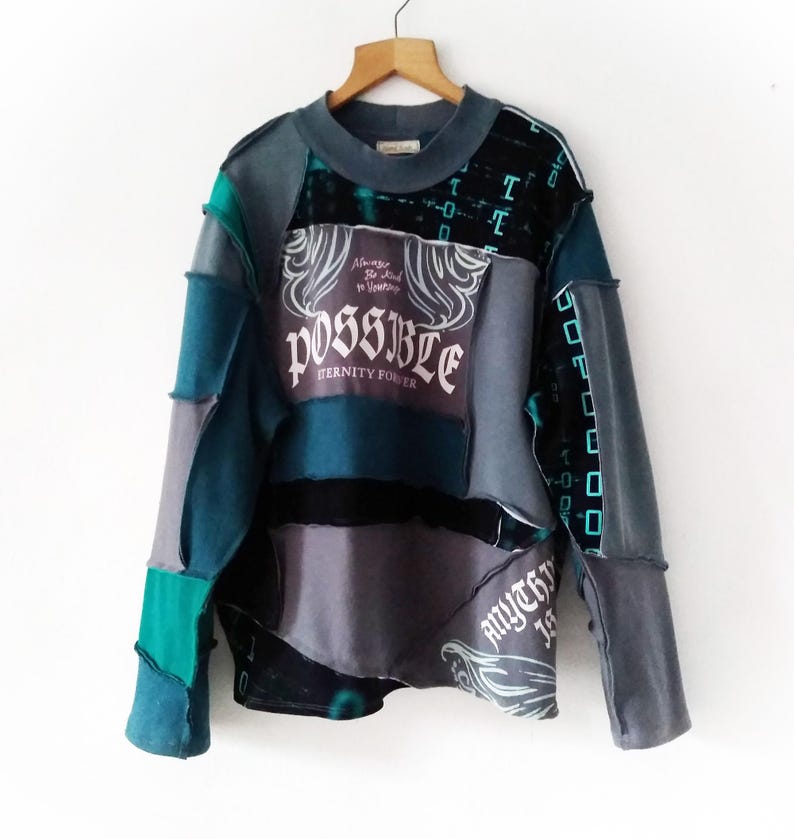 May include: A patchwork sweatshirt in shades of teal, gray, and black, hanging on a wooden hanger. The front features the word "POSSIBLE" with the text "Eternity Power" and "Always Be Kind to Yourself". The sleeves have a geometric design.