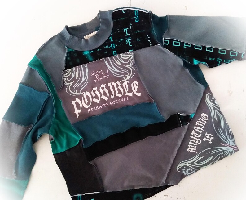 May include: A patchwork sweatshirt in shades of teal, gray, and black. The front features the word "POSSIBLE" in white script, with the phrases "Always Be Kind to Yourself" and "Eternity Forever." The sleeves and body are made of various fabric pieces.