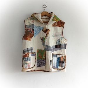 May include: A sleeveless hooded vest constructed from a patchwork of various fabrics including floral, plaid, paisley, and solid cream patterns. The garment features a front button closure, two large patch pockets, and brown trim along the edges.