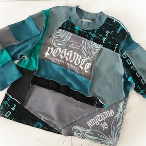 May include: A patchwork long-sleeve shirt in shades of gray, teal, and black. The shirt features the words "POSSIBLE Eternity Forever" and "ANYTHING IS" in white script. The design incorporates various fabric textures and patterns.