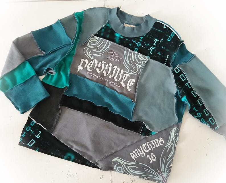 May include: A patchwork long-sleeve shirt in shades of gray, teal, and black. The shirt features the words "POSSIBLE Eternity Forever" and "ANYTHING IS" in white script. The design incorporates various fabric textures and patterns.