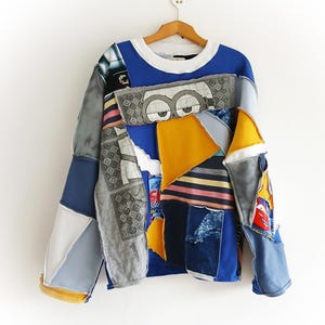 Może przedstawiać: Bluza typu patchwork z białym okrągłym dekoltem i niebieskimi rękawami. Korpus koszuli to kolaż różnych tkanin w odcieniach niebieskiego, szarego, żółtego i białego. Projekt zawiera postacie z kreskówek i geometryczne kształty.