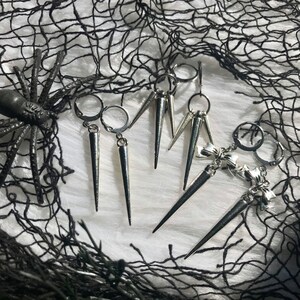 May include: A set of silver earrings with a variety of styles. One pair features a single spike, another pair has three spikes, and the last pair has a bow charm with a spike.