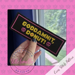 May include: A rectangular black cross-stitch piece featuring the yellow text "GODDAMNIT, DONUT!" and two small pink skull icons. The embroidery is bordered by a thin pink stitched line, creating a distinct contrast against the black fabric background.