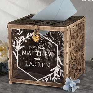 May include: A wooden wedding card box with intricate floral cutouts and a light blue envelope. The box features the names "MATTHEW and LAUREN" and the date "10.24.2026". A small gold lock with the word "Love" secures the box.