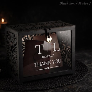 May include: Black, medium-sized box with intricate cut-out designs. The box has a clear front panel displaying a card with the initials "TL", the date "10.10.2027", and the words "THANK YOU" in white. A heart-shaped lock secures the lid.