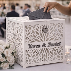 May include: A white, laser-cut wooden card box with the names "Karen & Daniel" in gray lettering. The box has a slot on top and a heart-shaped lock. A person is placing a gray envelope into the box. A bouquet of white roses is in the foreground.