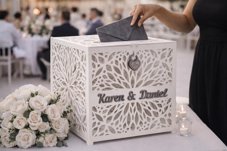 May include: A white, laser-cut wooden card box with the names "Karen & Daniel" in gray lettering. The box has a slot on top and a heart-shaped lock. A person is placing a gray envelope into the box. A bouquet of white roses is in the foreground.