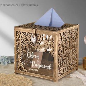 May include: A large square wooden card box with an old wood finish and intricate laser-cut willow branch patterns. The front features a clear panel with the white printed text Coleen & Raymond and 5/30/2026. A heart-shaped padlock secures the top lid.