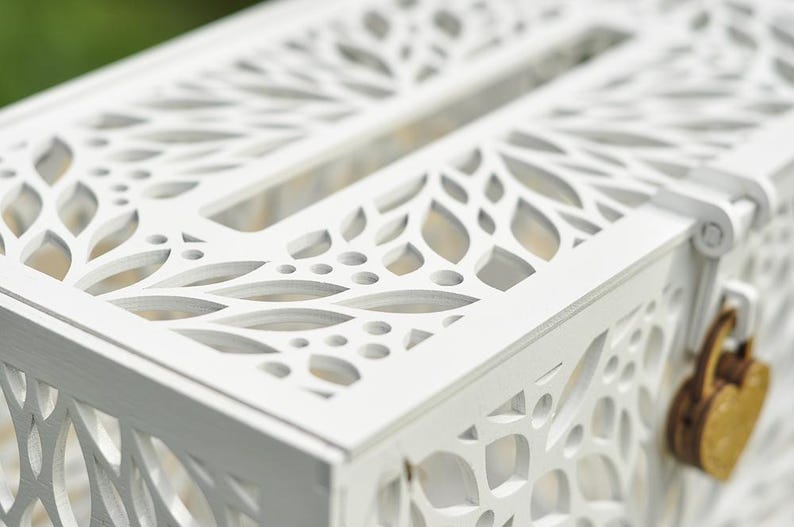 May include: A white wooden card box with intricate leaf-shaped cutouts. The box has a slot on top and a heart-shaped lock. The design is detailed and elegant, suitable for weddings or special events.