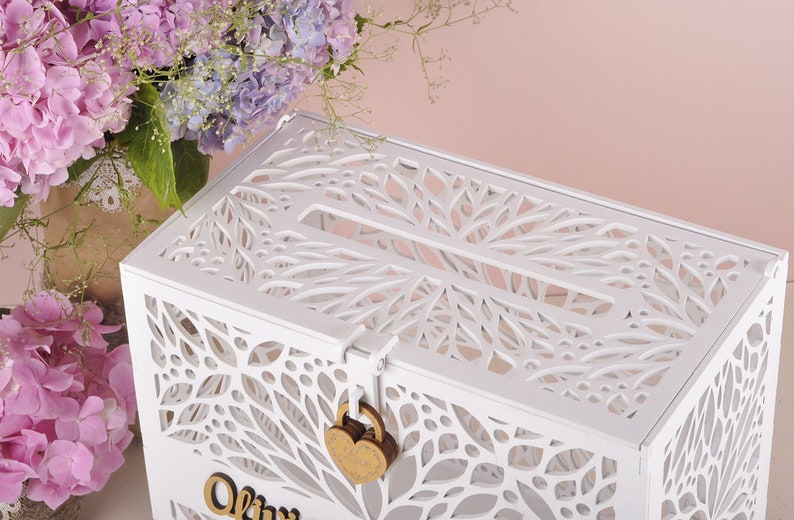 May include: A white wooden card box with a decorative leaf pattern cut into the sides and top. The box has a slot on top and a heart-shaped lock. Pink and purple flowers are in the background.