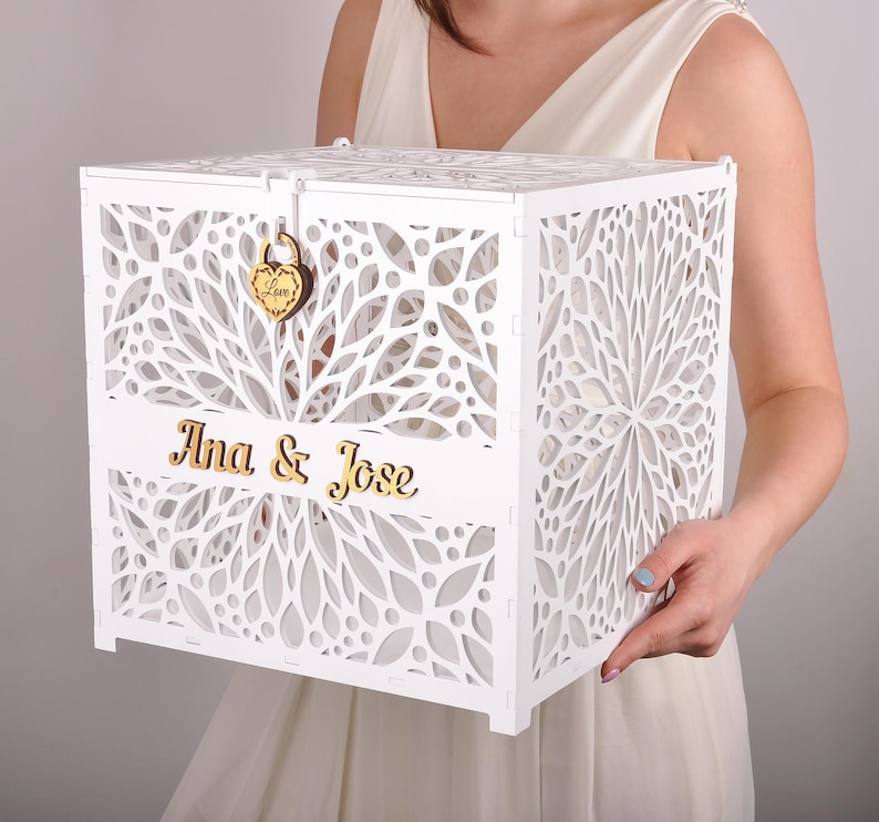 May include: A white, laser-cut wooden box with floral patterns and the names "Ana & Jose" in gold lettering. The box has a heart-shaped lock with the word "Love" engraved on it. Ideal for weddings or special events.